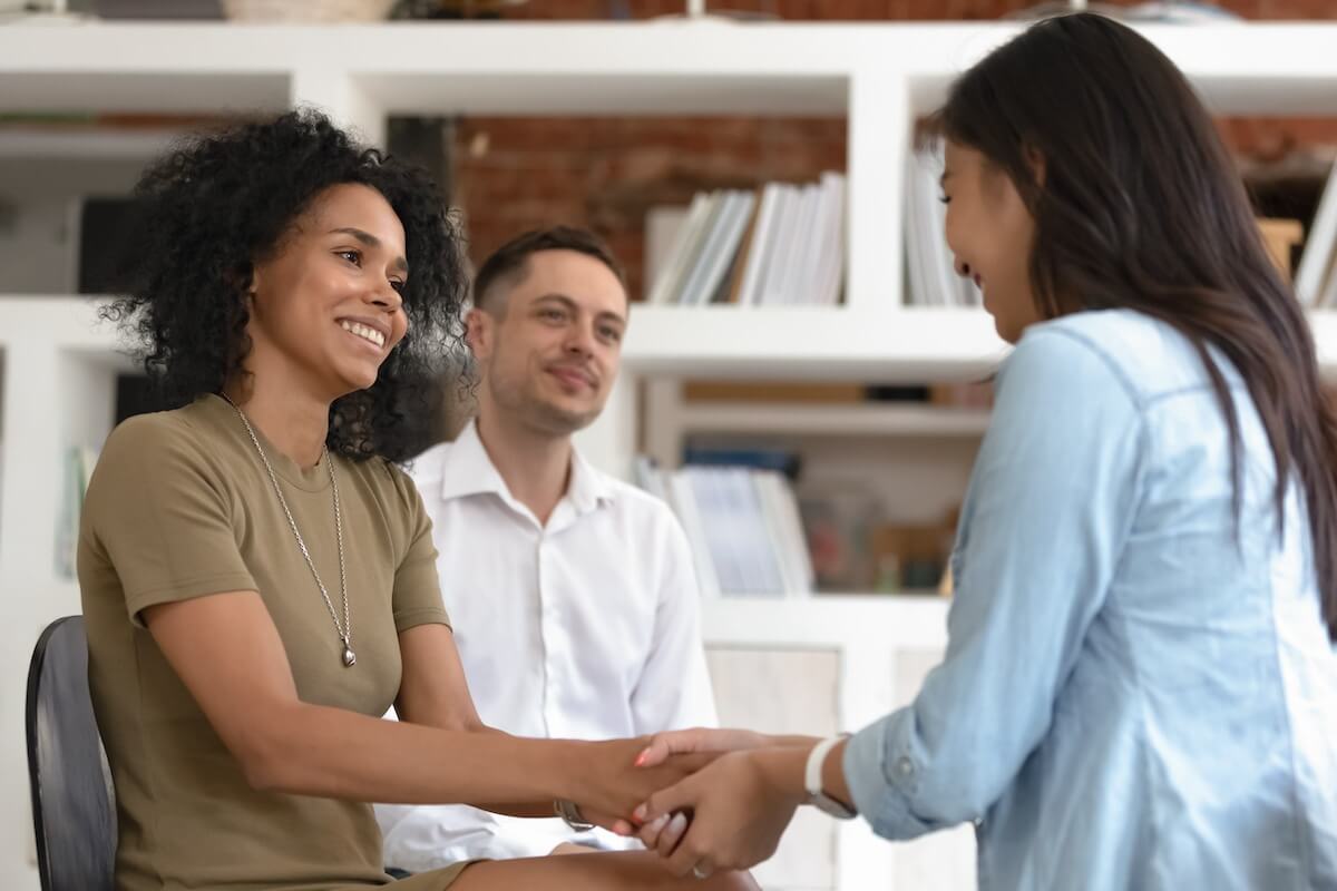 Two women shake hands in a professional setting. Nearby, a man sits smiling.