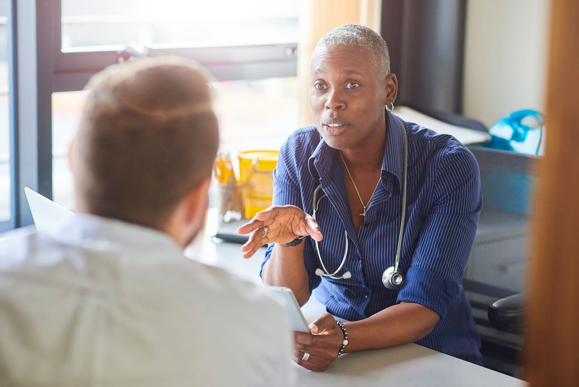 A female doctor sits at her desk and chats to a male patient while looking at his test results on her digital tablet . She is a blue shirt with the sleeves rolled up and a stethoscope around her neck.